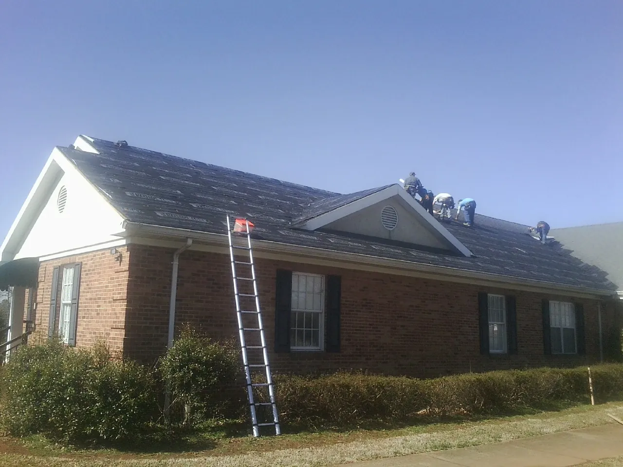 Crew tearing shingles off a brick ranch roof with a ladder set — Emergency Roof Repair work by our crew in Bassett