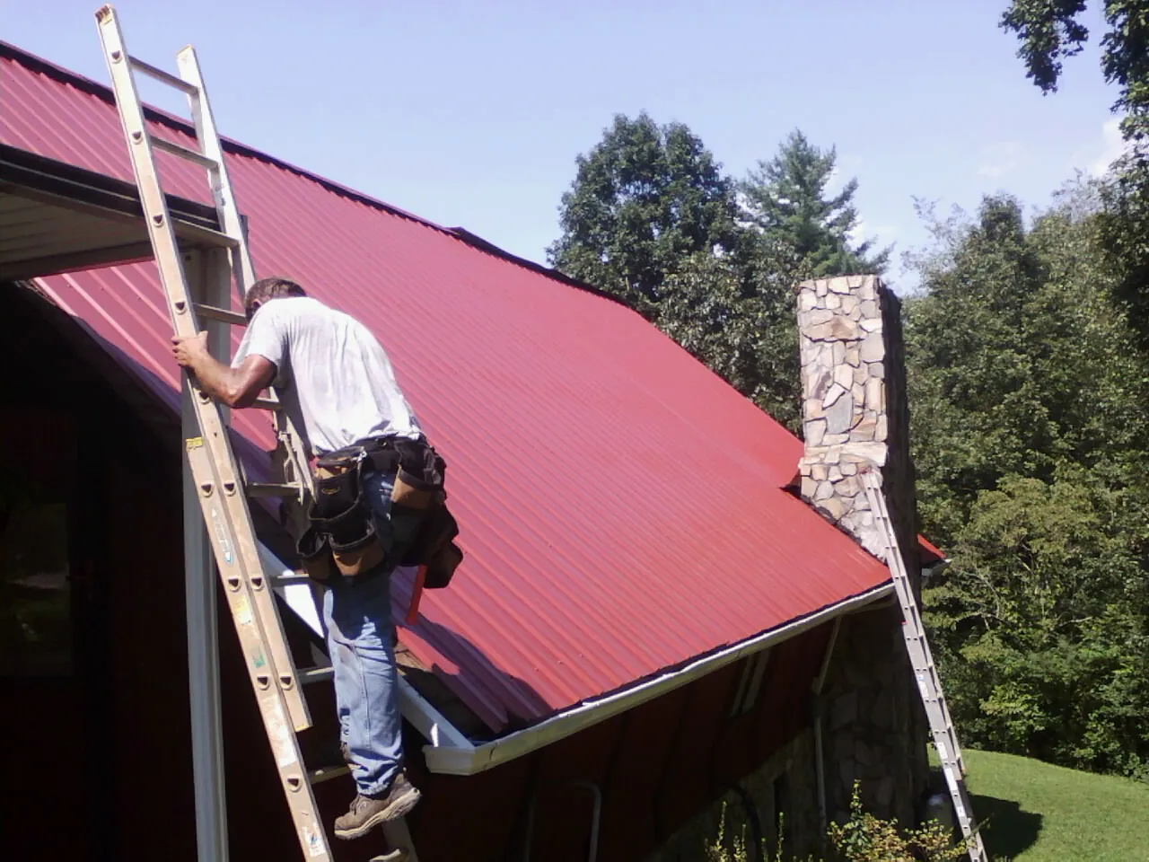 Roofer on a ladder beside a red metal roof and stone chimney — Soffit & Fascia Repair by Roofing Brothers in Bassett
